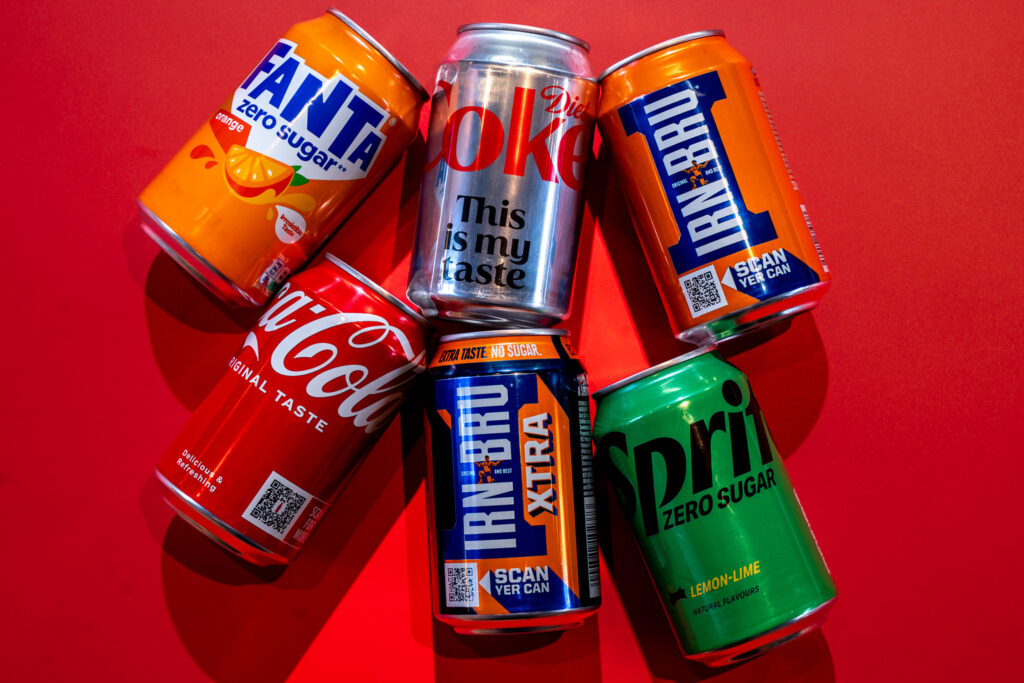 selection of fanta, coca-cola, diet coke, irn-bru, irn-bru extra and sprite cans lying on red background