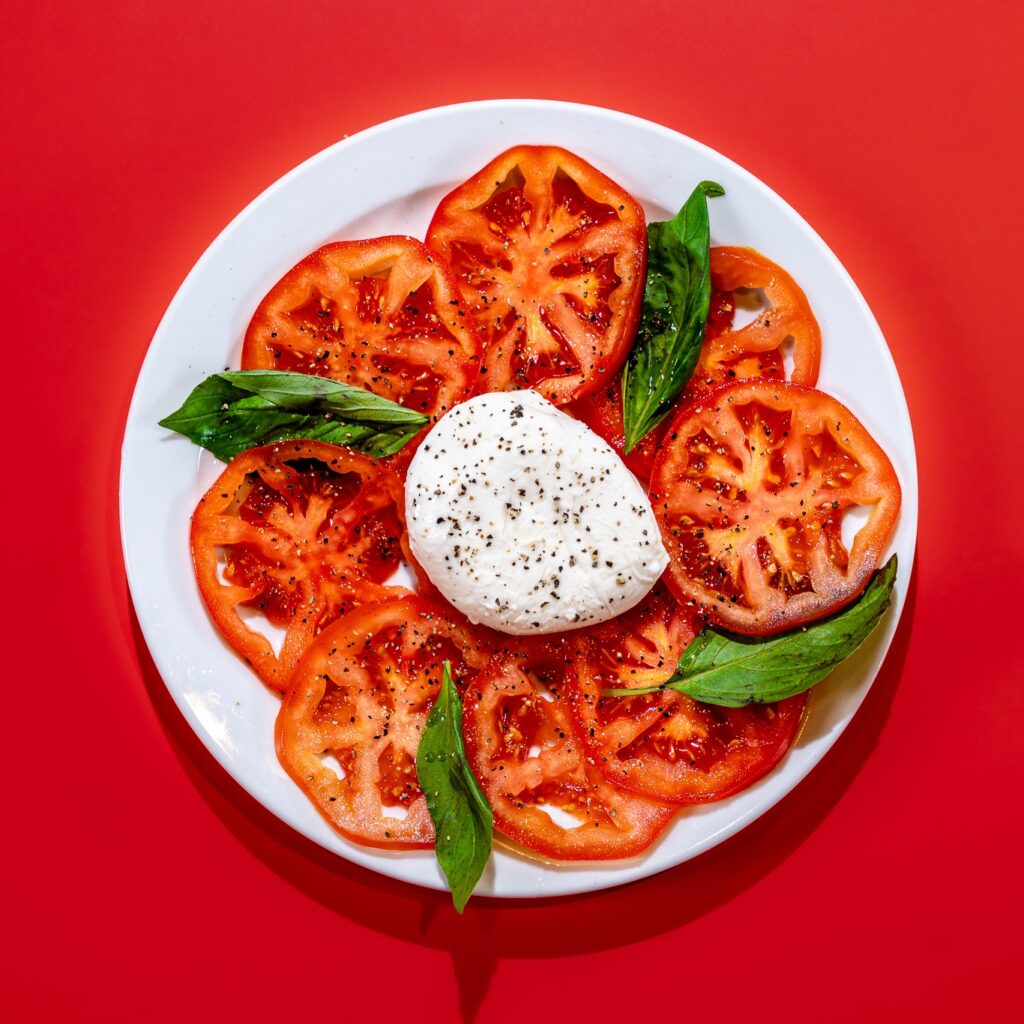 sliced tomatoes, basil and whole buffalo mozzarella on white plate with red background